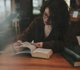 A person sits at a wooden table in a dimly lit room, reading an open book with another large book resting nearby