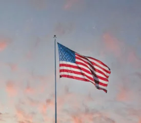 An American flag waves on a tall pole against a pastel evening sky with soft pink clouds