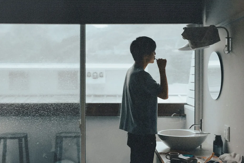 Man brushing their teeth in a small bathroom with a large window overlooking a rainy landscape