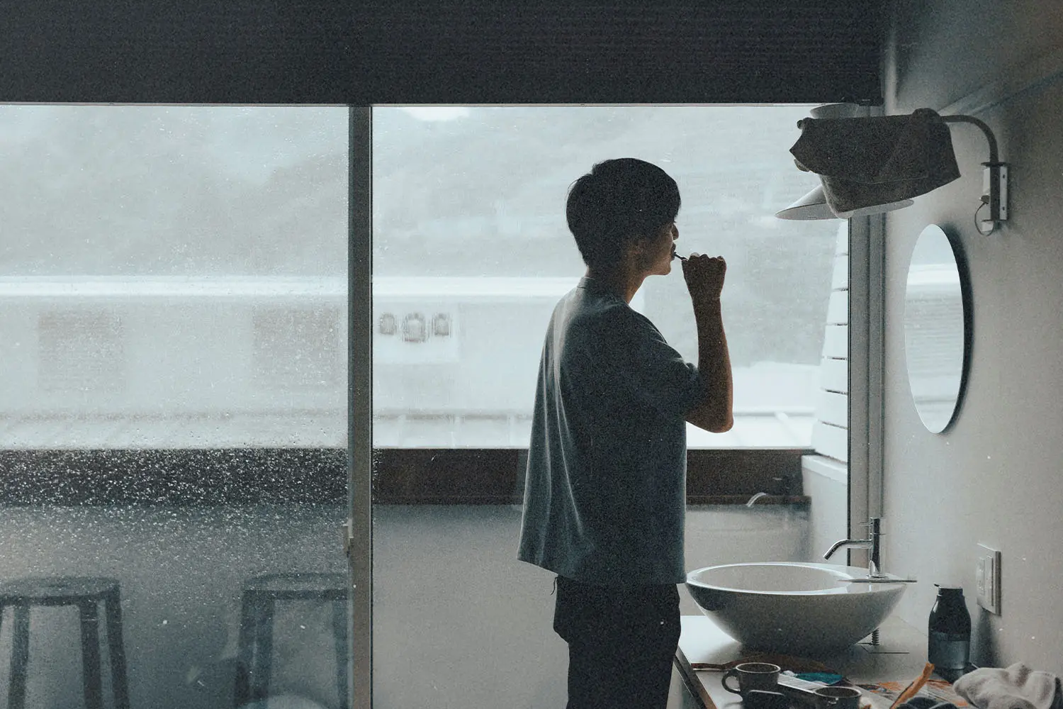 Man brushing their teeth in a small bathroom with a large window overlooking a rainy landscape