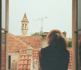 Person stands at an open window looking out over terracotta rooftops and a stone bell tower in a historic town