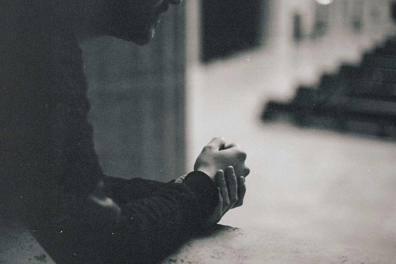 A person leans forward against a concrete ledge with hands clasped together, looking out over a quiet, empty walkway in a muted indoor setting