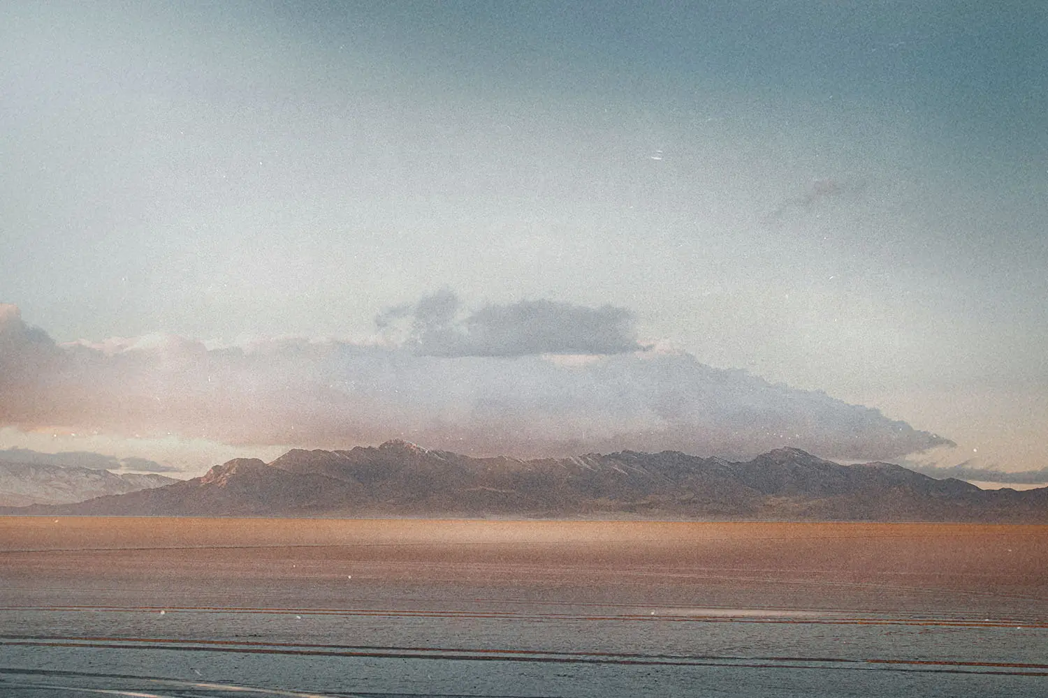 A wide, tranquil landscape showing a flat body of water in the foreground and a low mountain range beneath a soft, pastel-colored sky