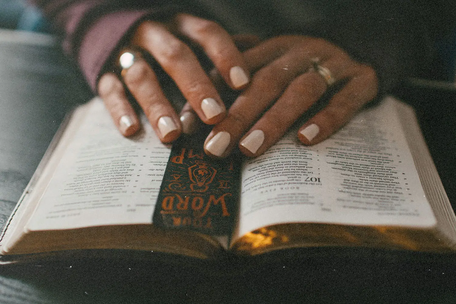 Hands rest on an open Bible with a decorative bookmark placed between the pages, shown in close-up