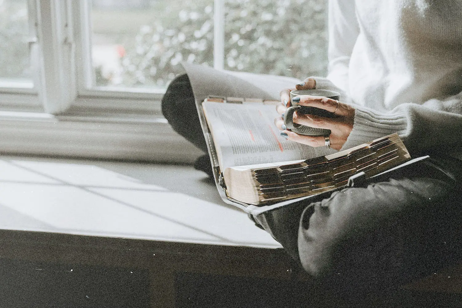 A person sits by a window holding a mug, with an open Bible resting on their lap as natural light fills the space