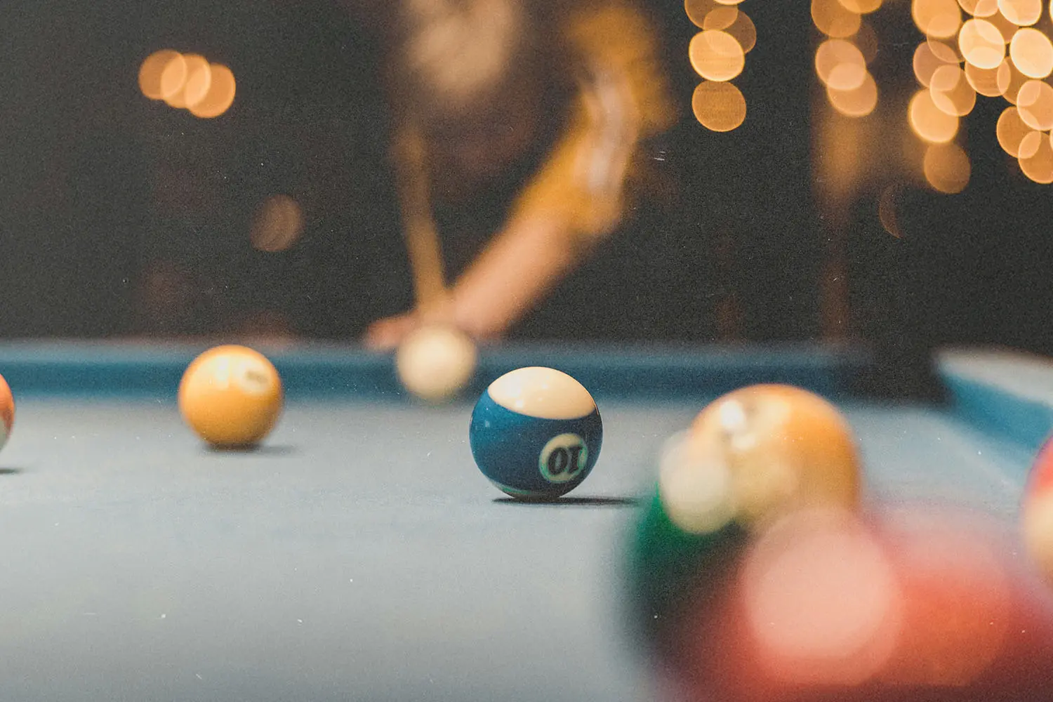 Billiard balls arranged on a pool table, with one ball centered in focus while a cue and other balls sit in the background under warm lighting