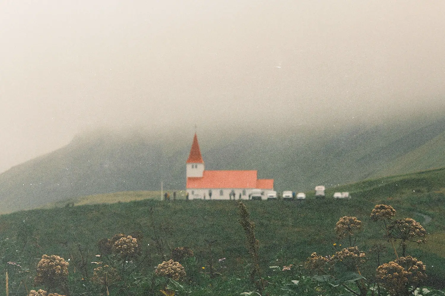 A small church with a red roof standing alone in a grassy field, surrounded by misty hills and low clouds