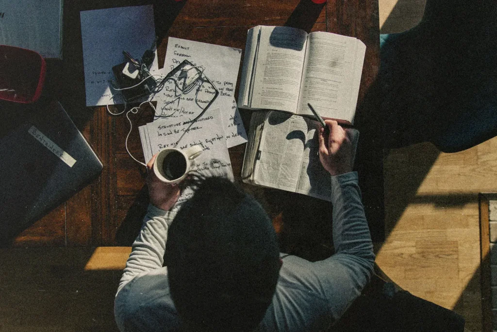 A person sits at a wooden table studying, surrounded by an open Bible, handwritten notes, earphones, and a cup of coffee
