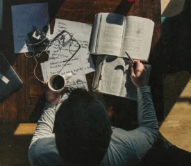 A person sits at a wooden table studying, surrounded by an open Bible, handwritten notes, earphones, and a cup of coffee