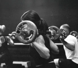 A group fitness class performs barbell squats, with participants lifting weighted bars across their shoulders