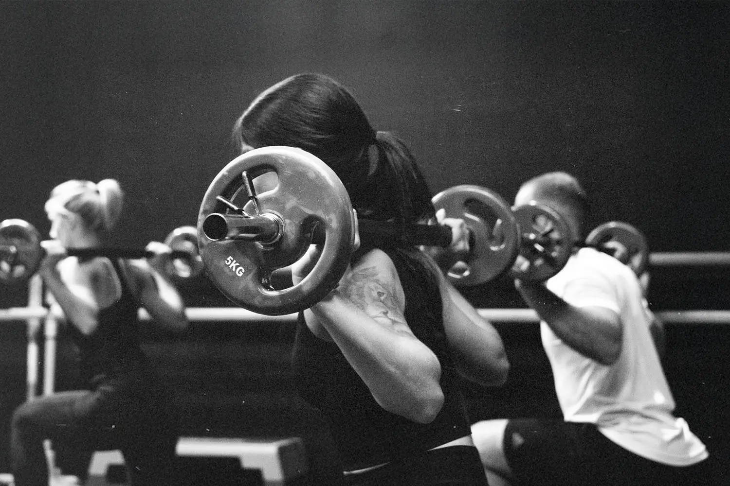 A group fitness class performs barbell squats, with participants lifting weighted bars across their shoulders