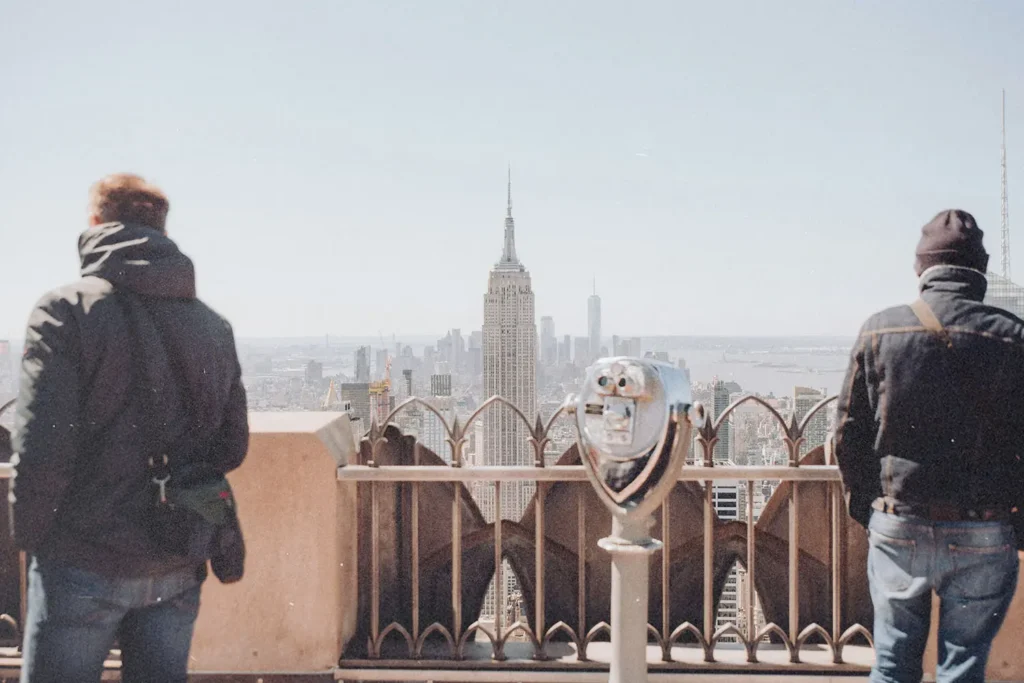 Two people standing at an observation deck overlooking the New York City skyline, with the Empire State Building centered in the view and a coin‑operated viewer in the foreground