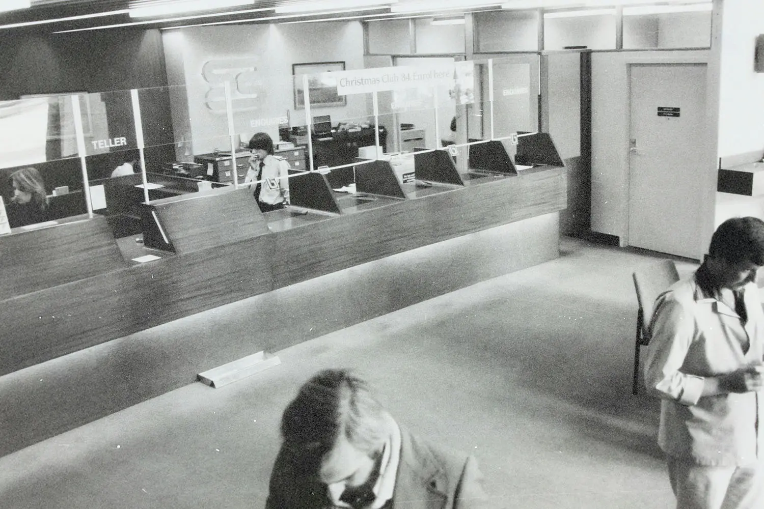 Interior of a bank lobby with teller counters, glass partitions, and several people moving through the space