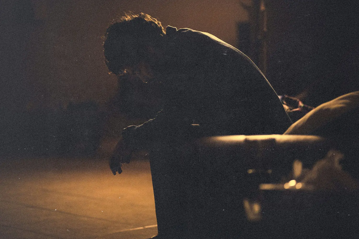 A person sits leaning forward on a couch in a dimly lit room, illuminated by warm, low light