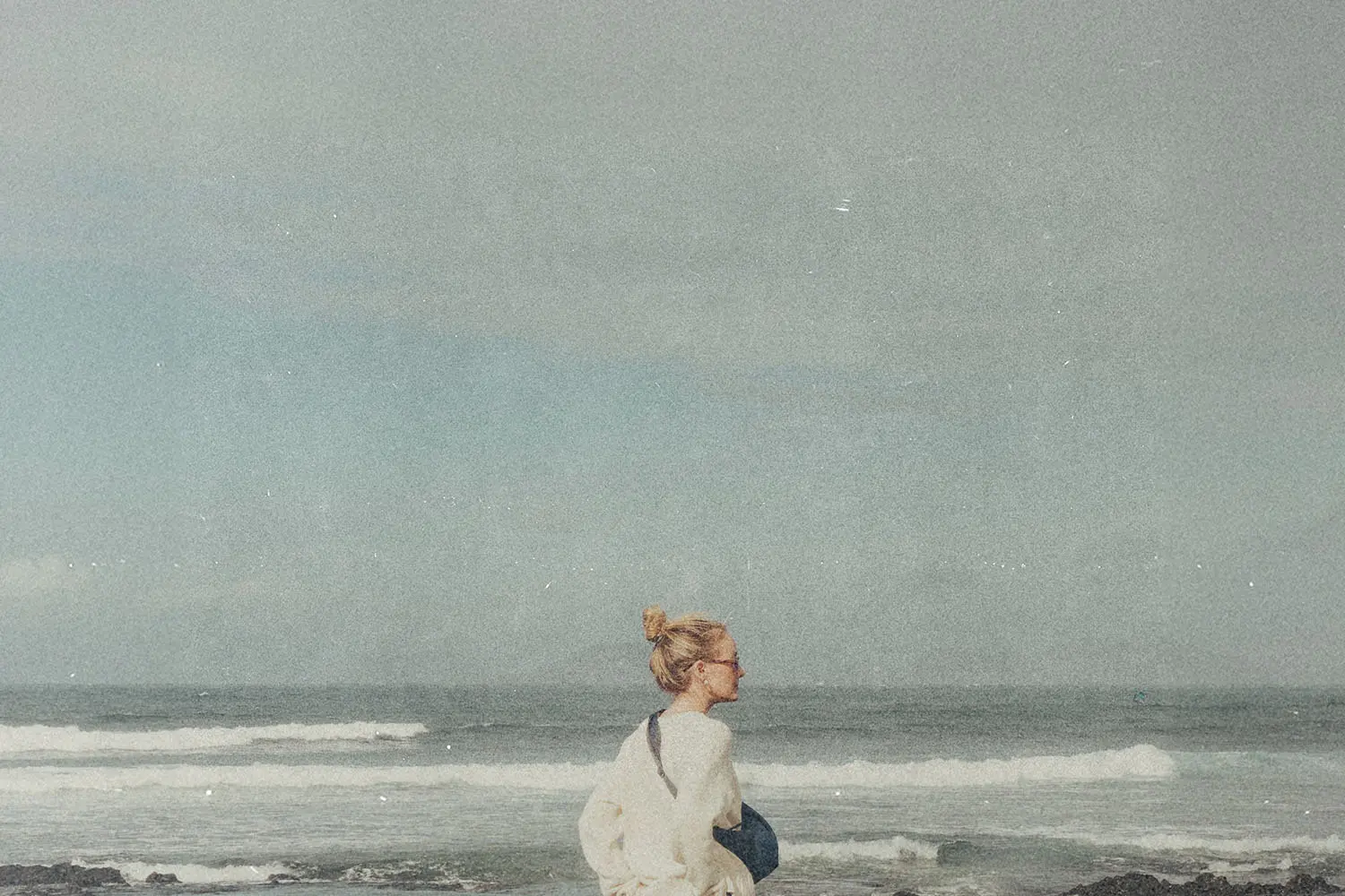 A person stands near the shoreline, facing the ocean as waves roll in under a pale sky