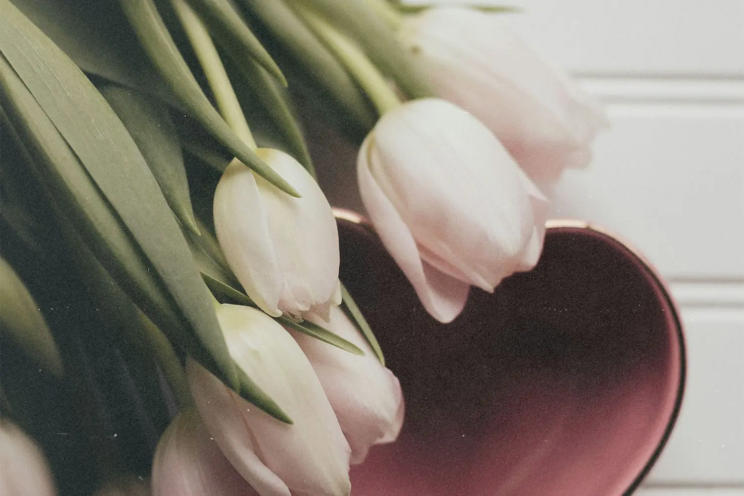 White tulips spill from a vase beside a heart-shaped dish on a light-colored surface
