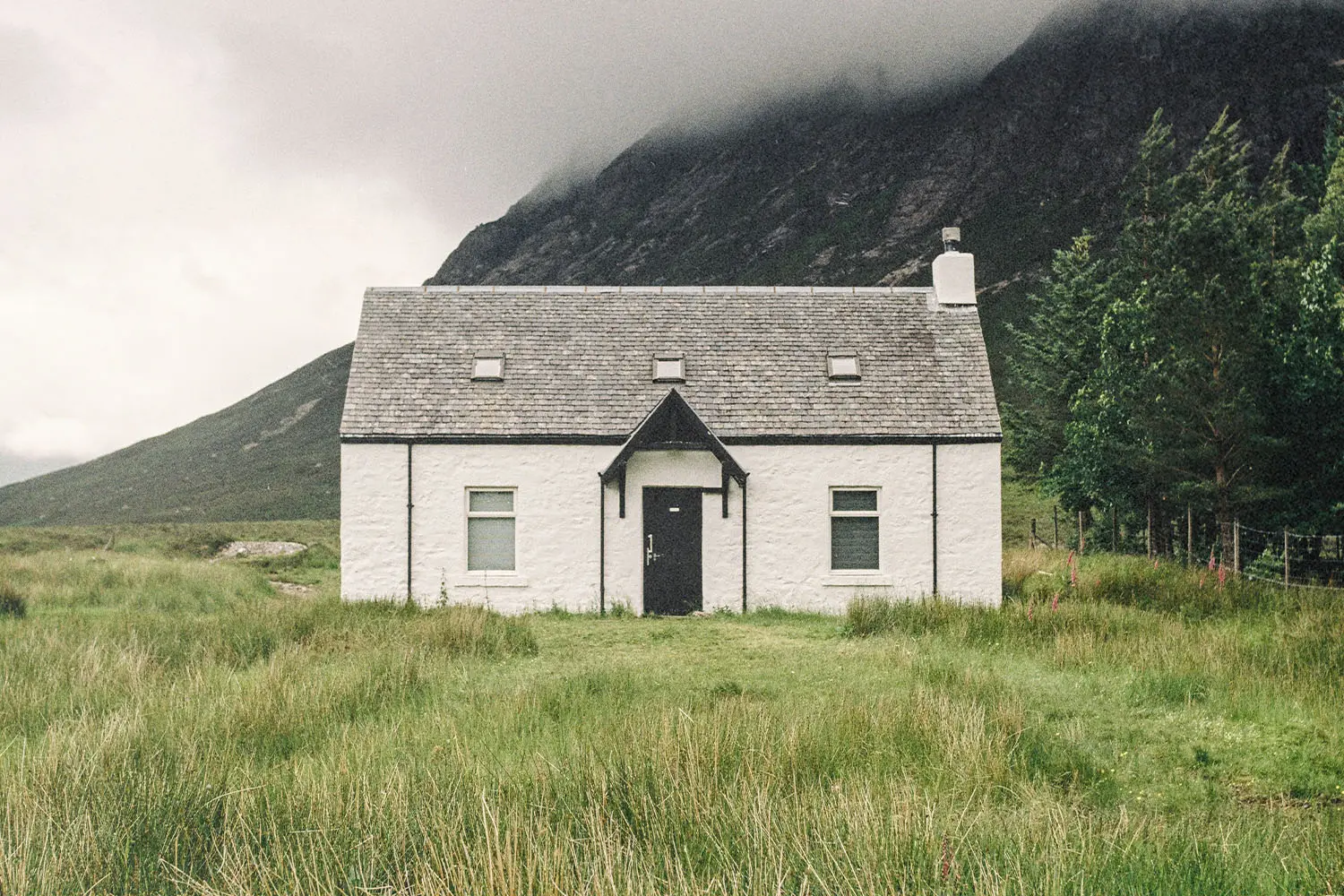 A small white house stands alone in a grassy field with misty mountains rising behind it
