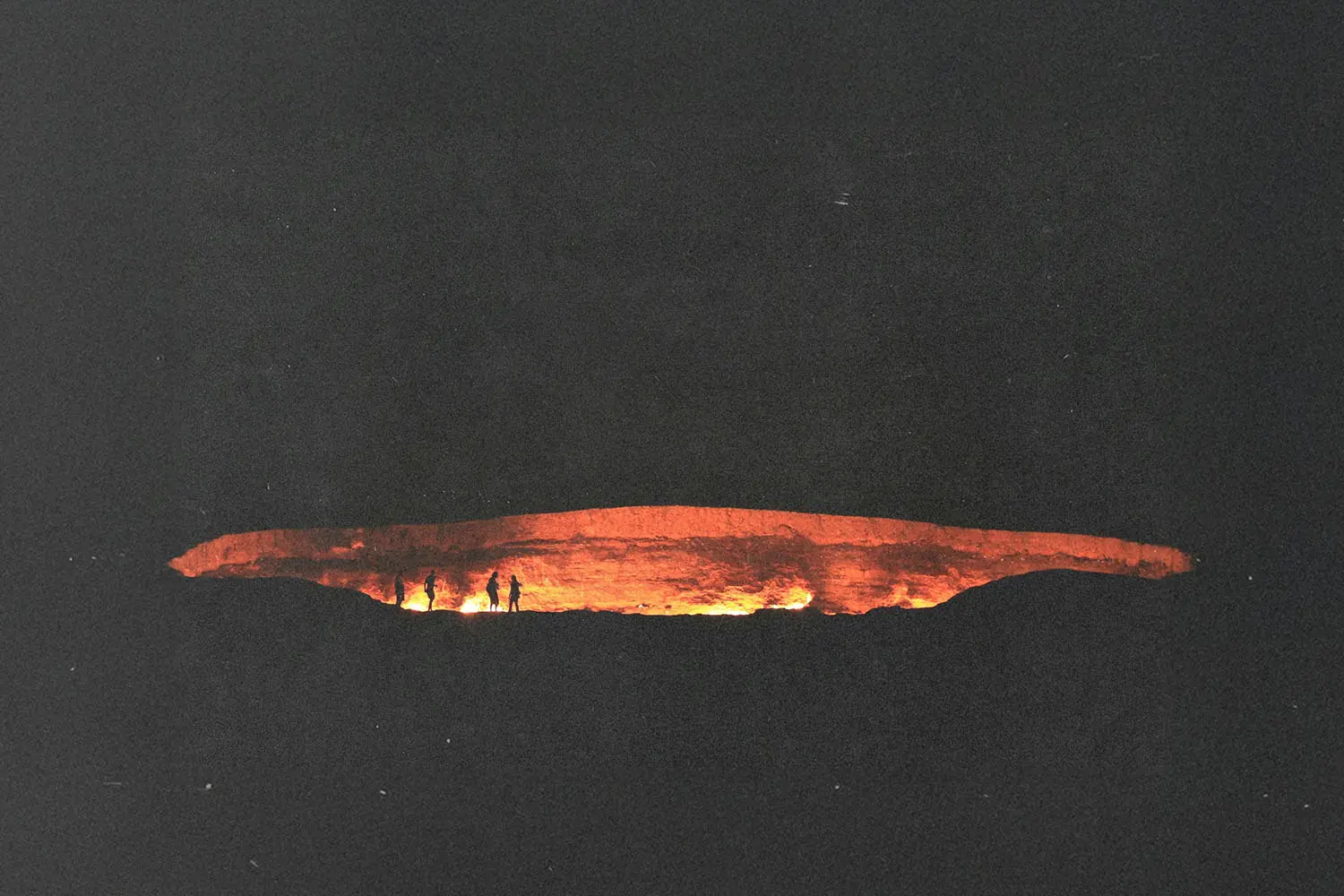 A wide view of a glowing crater at night with several small figures standing along the edge