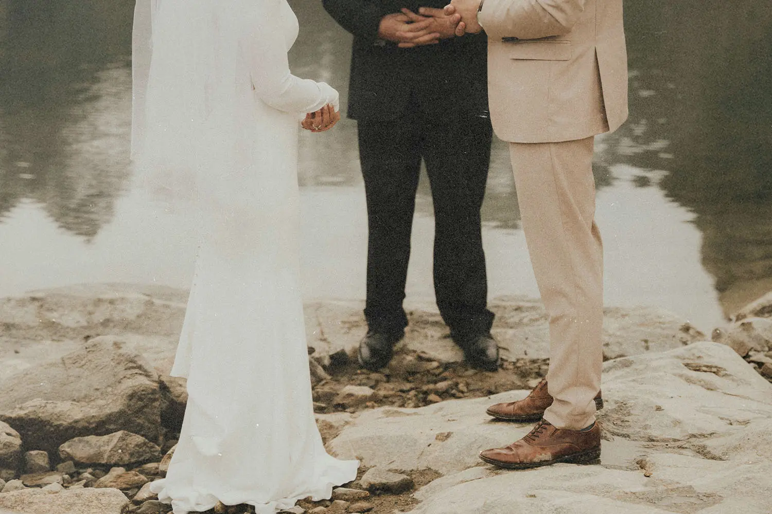 Two people standing on rocky ground beside calm water during a wedding ceremony, with an officiant positioned between them