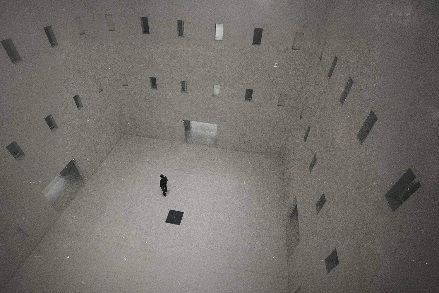 A solitary person standing in the center of a large, minimalist concrete interior with tall walls and narrow windows