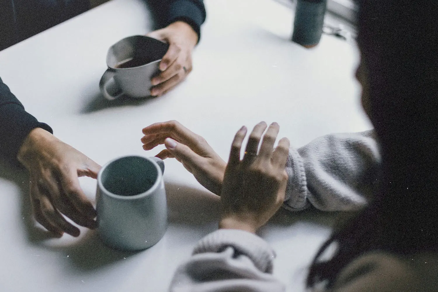Three hands resting near ceramic mugs on a white tabletop, suggesting conversation over drinks