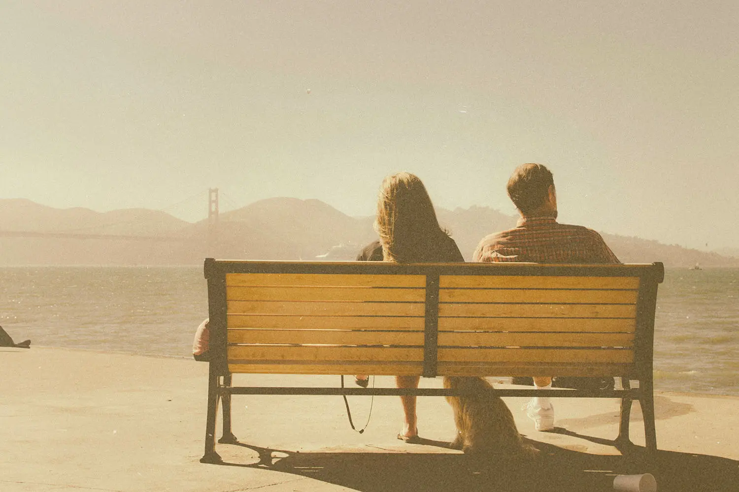 Two people seated on a wooden bench facing a body of water, with distant hills and a bridge visible on the horizon