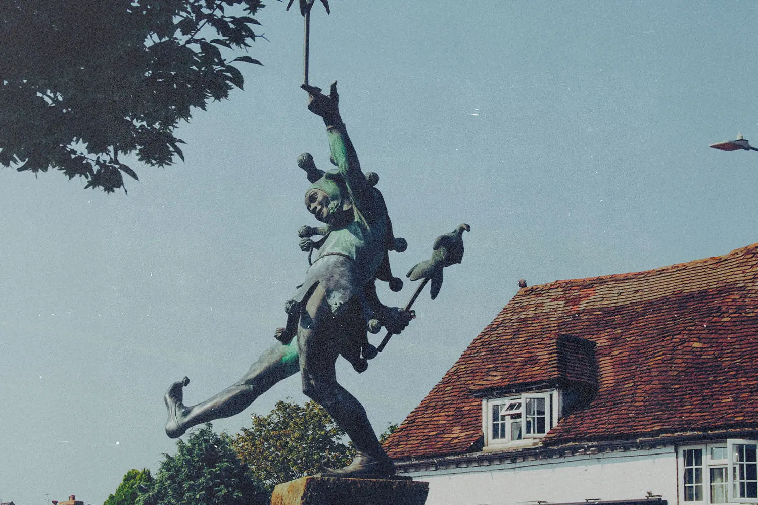 A bronze statue of a lively, dancing figure holding a staff stands in front of a historic building with a red‑tiled roof