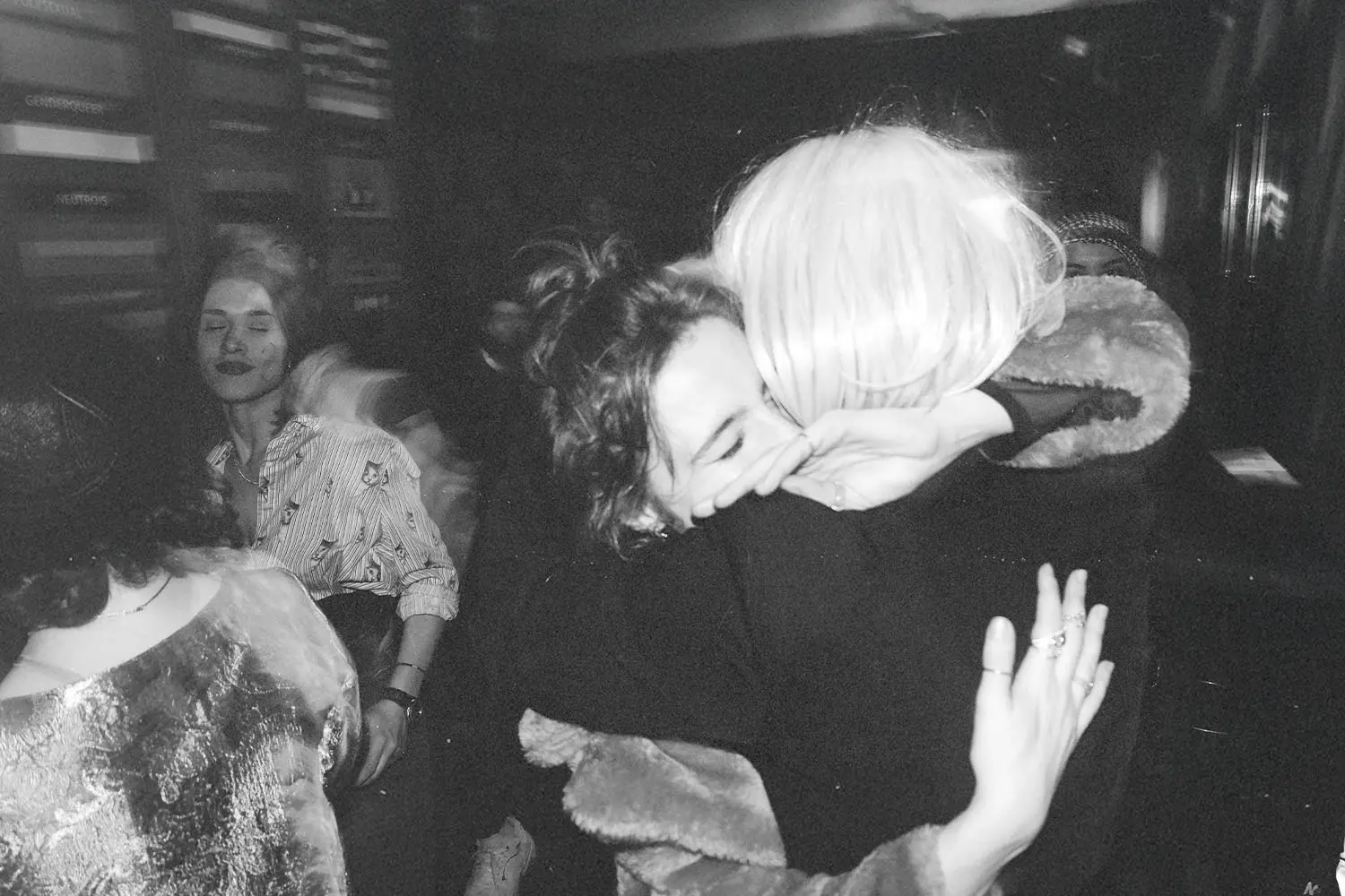 Two people embracing tightly in a crowded indoor space with others gathered and talking in the background