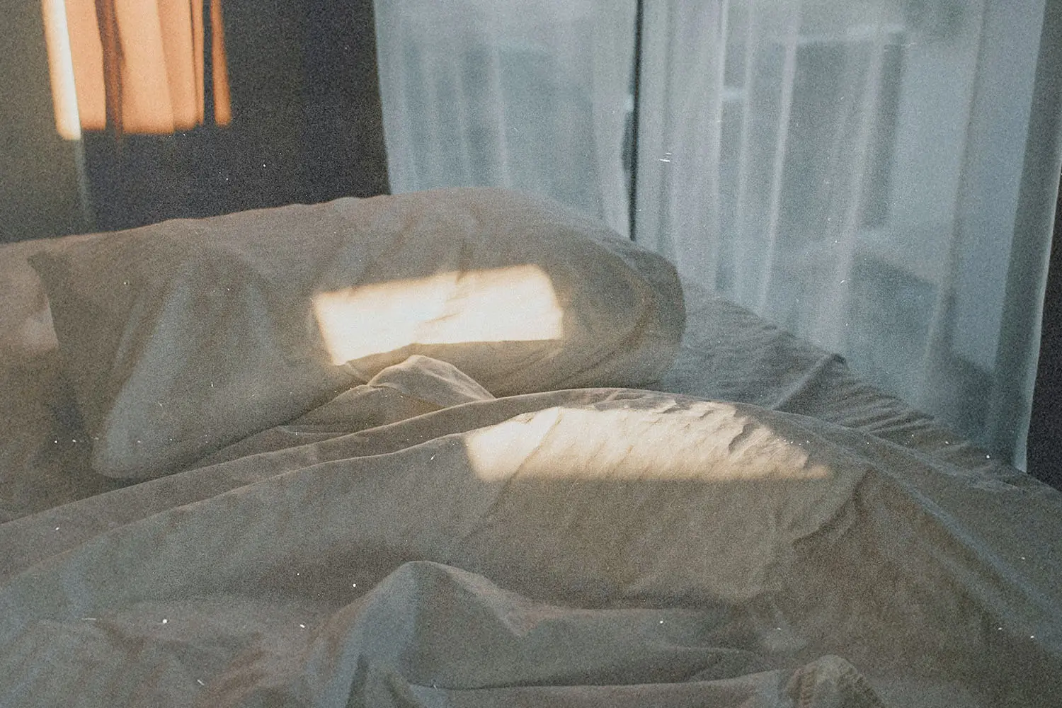 Soft morning light shining across rumpled bedding and pillows near a window with sheer curtains
