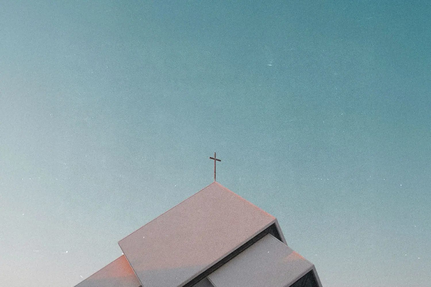 Low-angle view of a modern church roof with angular lines and a cross at the peak, set against a clear blue sky with subtle film grain texture