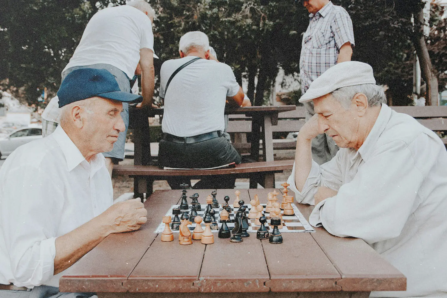 Two elderly males sit across from each other at a wooden picnic table outdoors playing a game of chess
