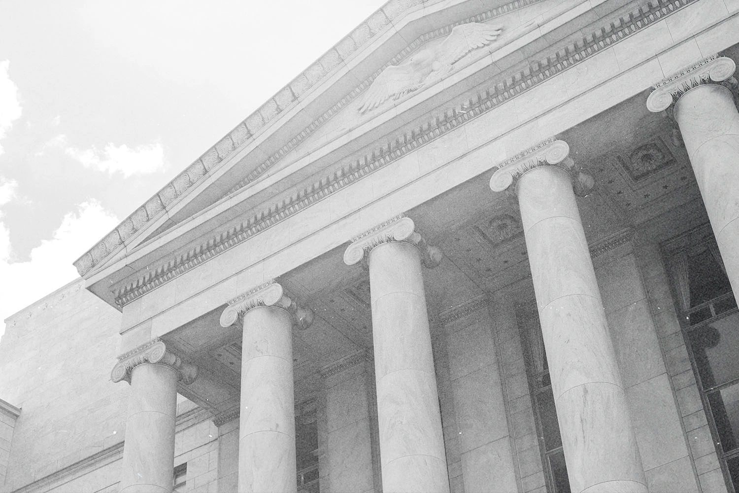Low-angle black-and-white view of a large neoclassical building with tall columns and a triangular pediment against a cloudy sky