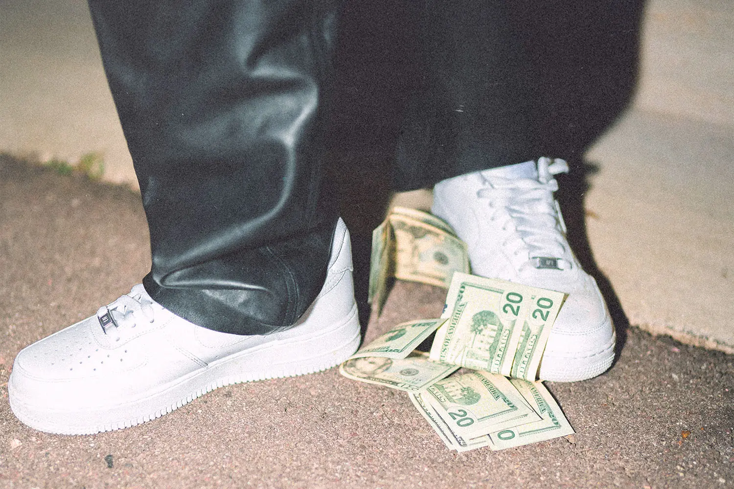 White sneakers on pavement next to several U.S. dollar bills scattered near the ground