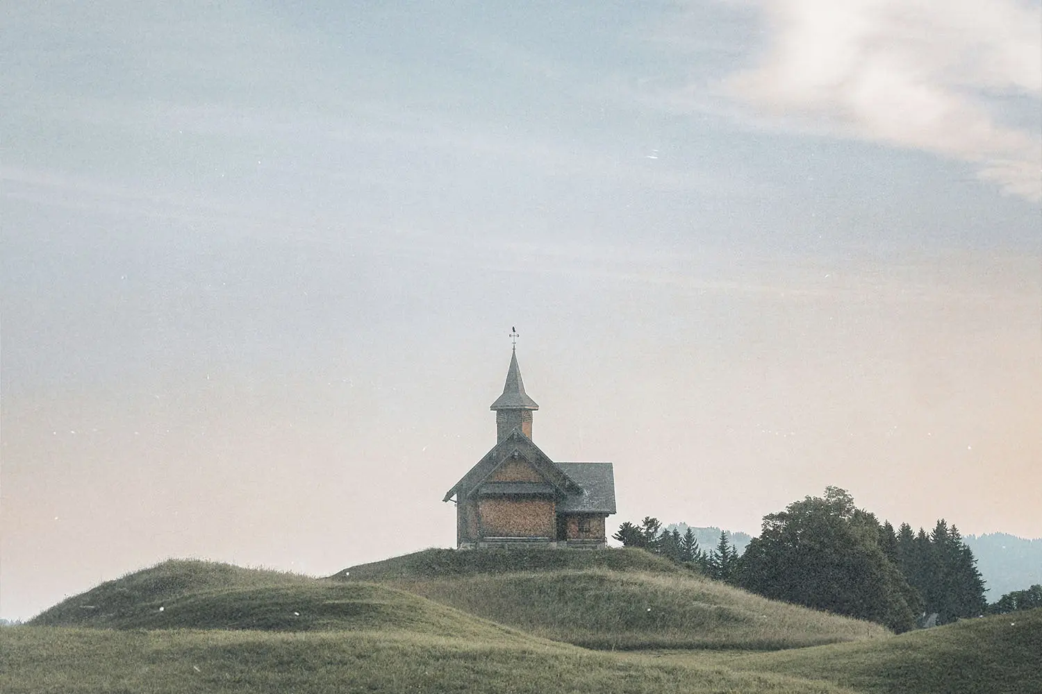 A small wooden church sits alone on a grassy hill beneath a wide, pale blue sky