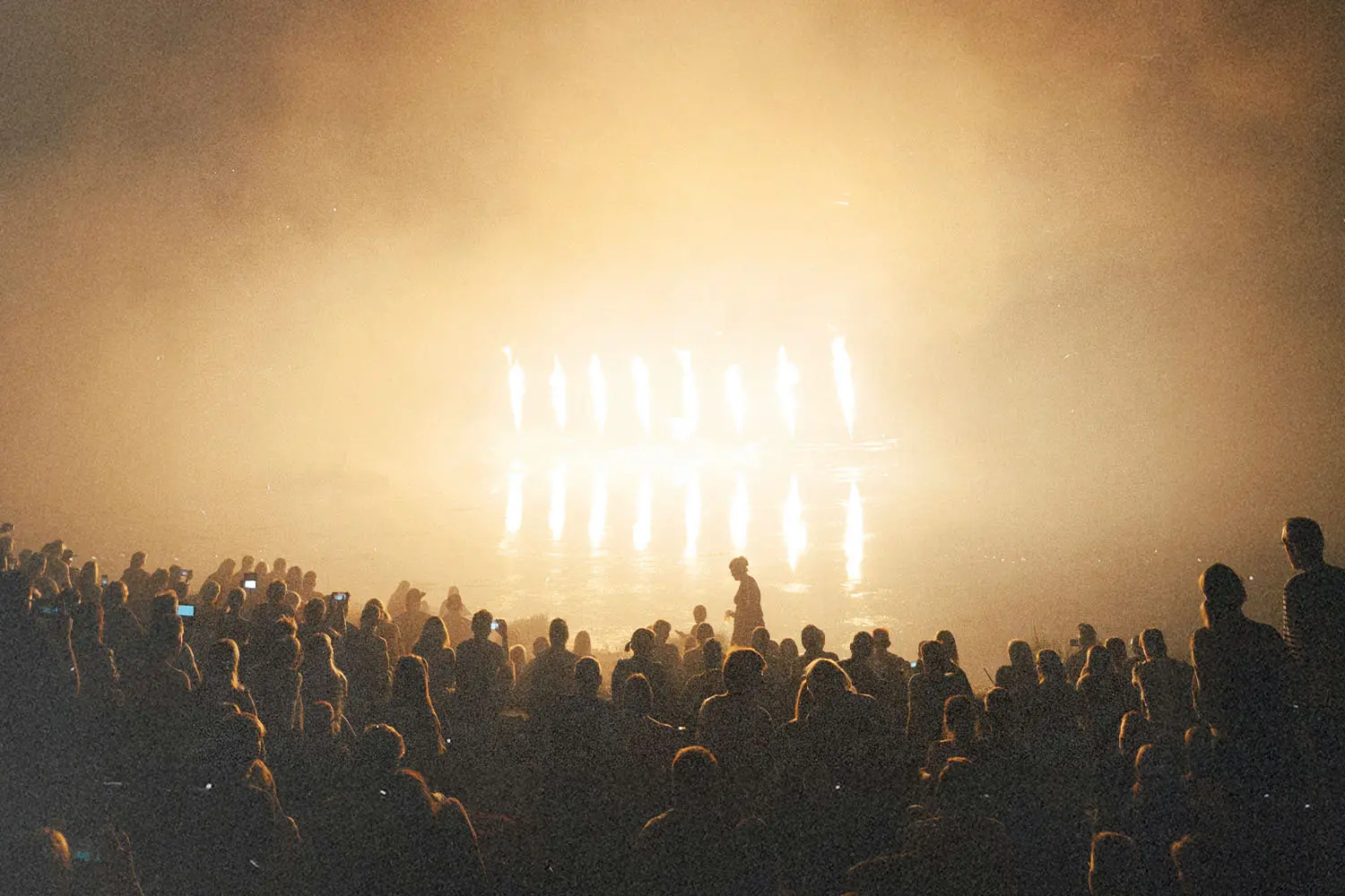 A large crowd gathered indoors, silhouetted against intense golden stage lights shining through haze