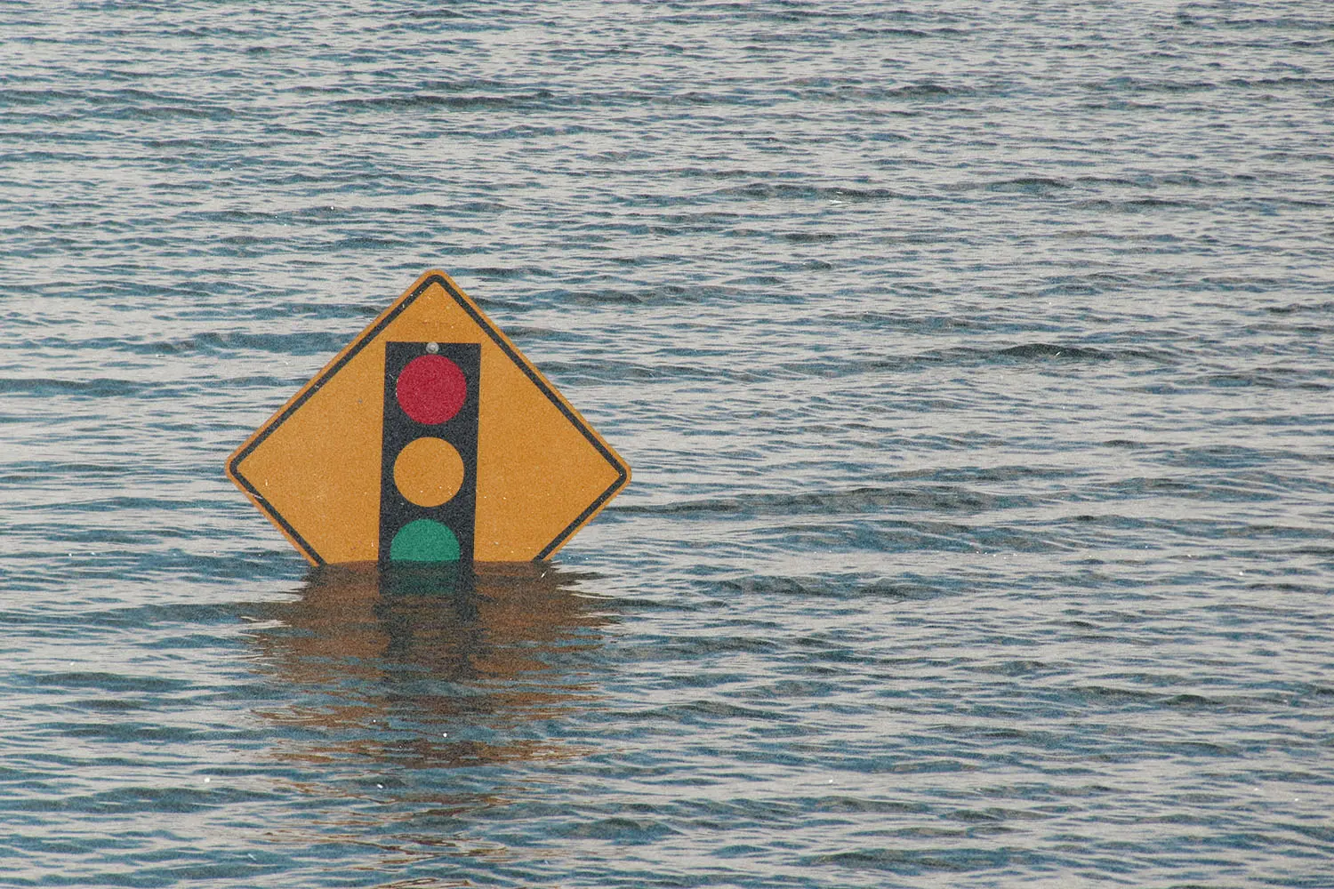 A traffic signal warning sign partially submerged in floodwater, with rippling water stretching across the scene