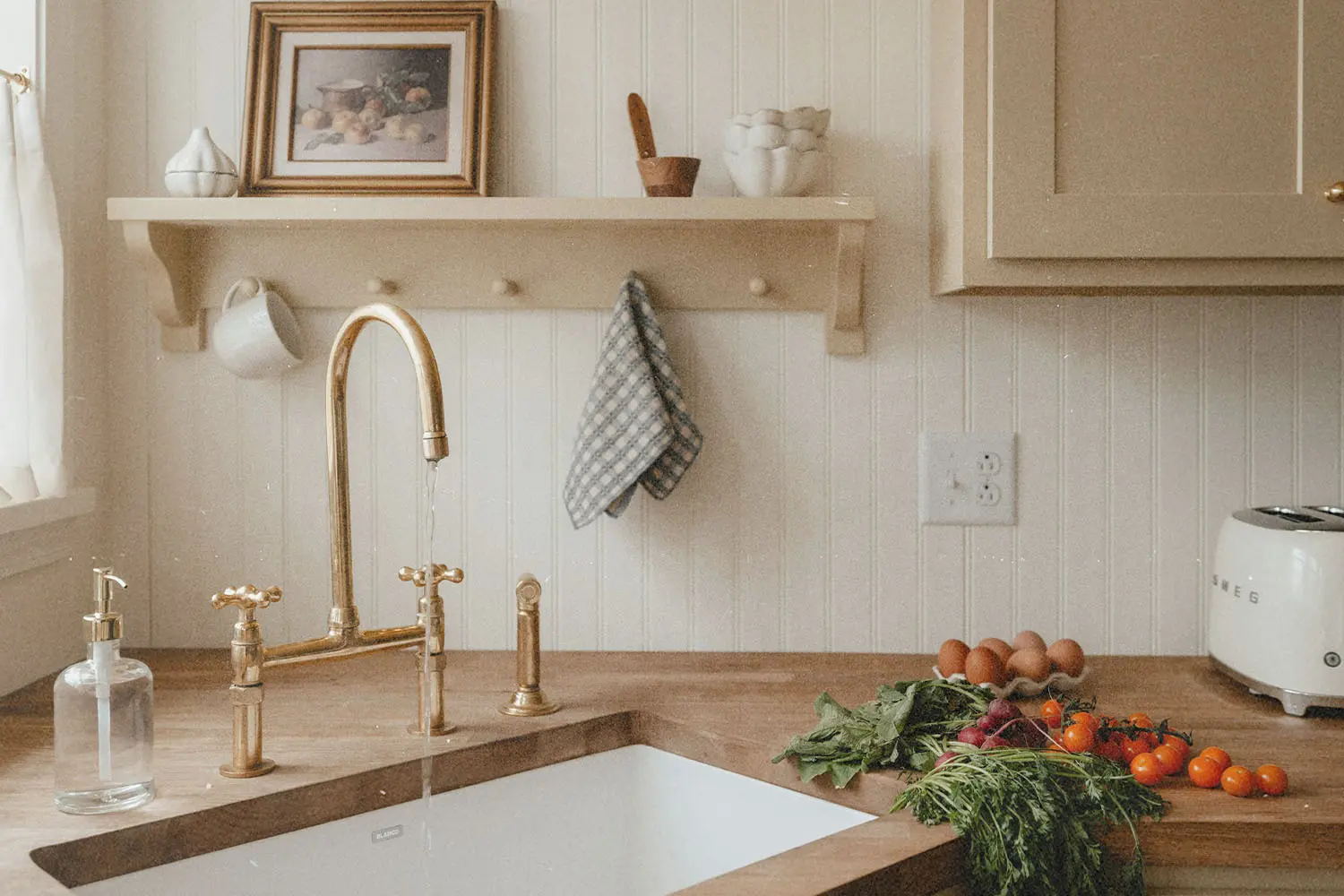 A bright, cozy kitchen sink area with a brass faucet, wooden countertop, fresh vegetables, eggs, and shelves holding simple decor