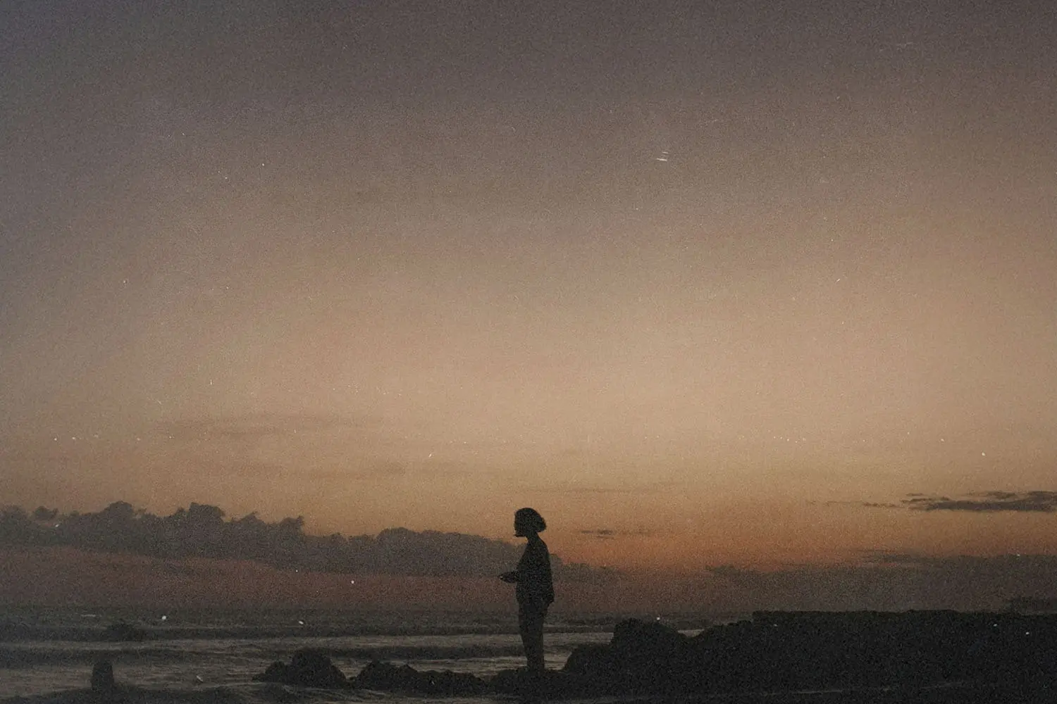 A single person standing on rocky ground by the ocean at sunset, silhouetted against a softly colored sky