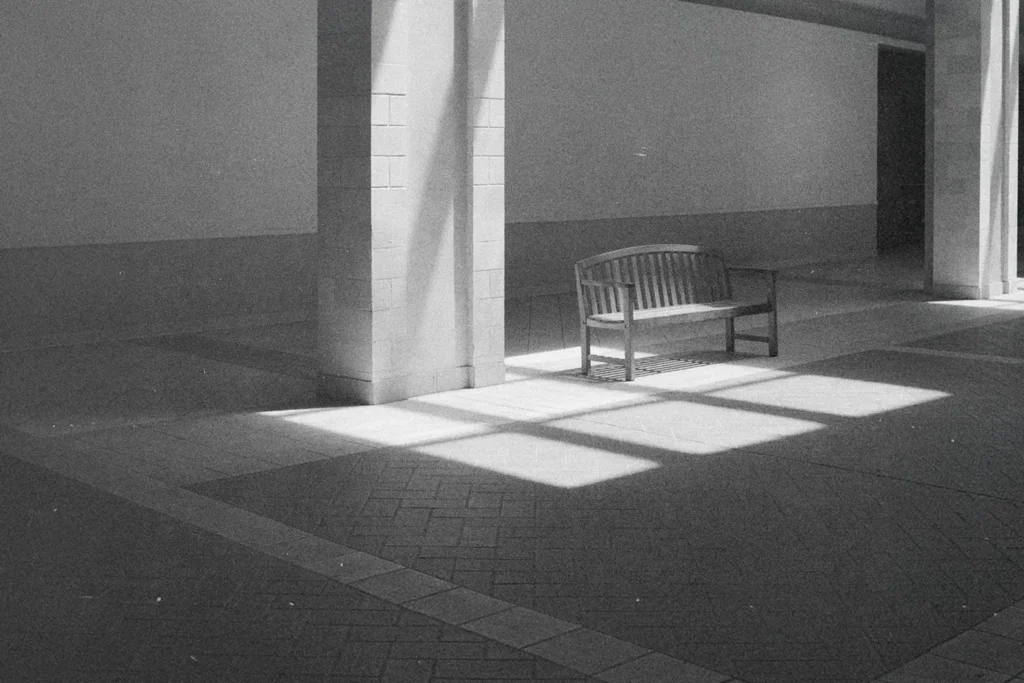 Sunlight shines through tall windows onto an empty wooden bench in a quiet indoor hallway with tiled floors