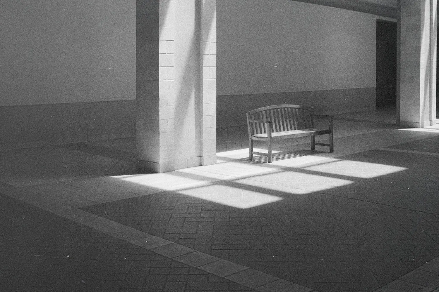 Sunlight shines through tall windows onto an empty wooden bench in a quiet indoor hallway with tiled floors