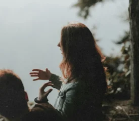 Two people sitting near the edge of a wooded area beside calm water, with one person gesturing while speaking