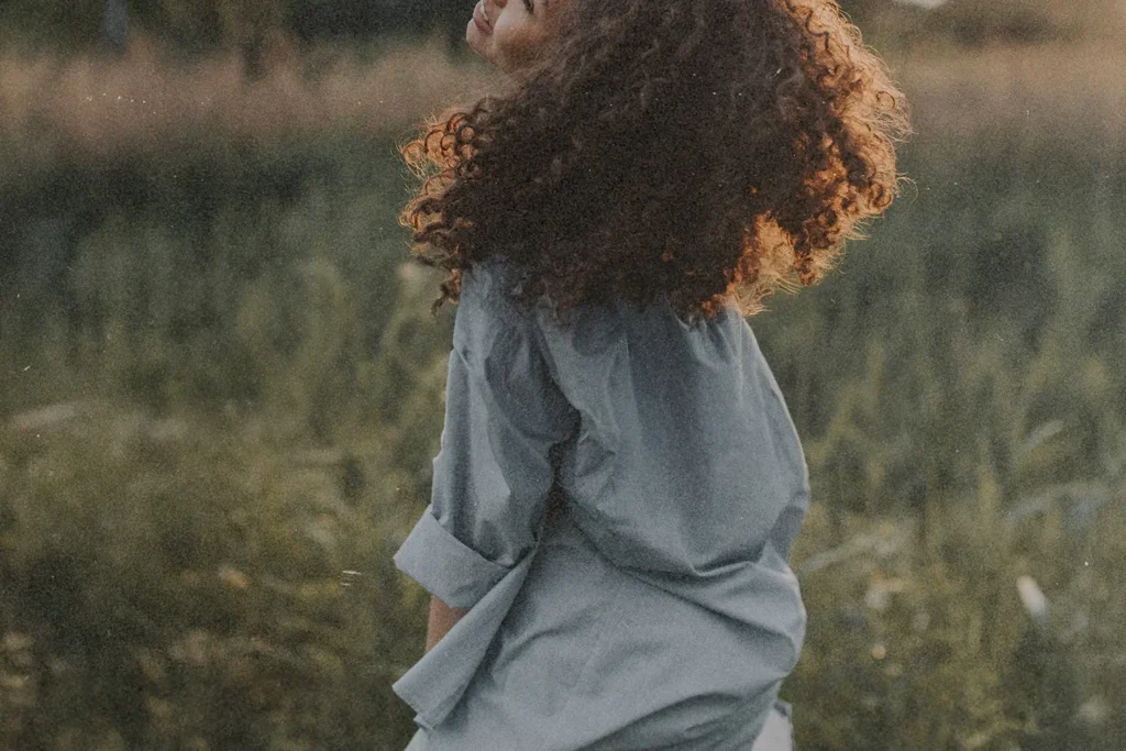 Person with curly hair standing in a grassy field with warm sunlight shining from behind
