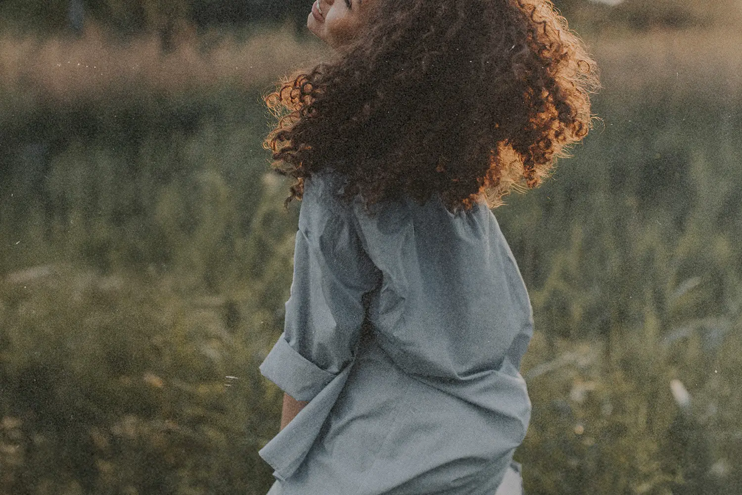 Person with curly hair standing in a grassy field with warm sunlight shining from behind