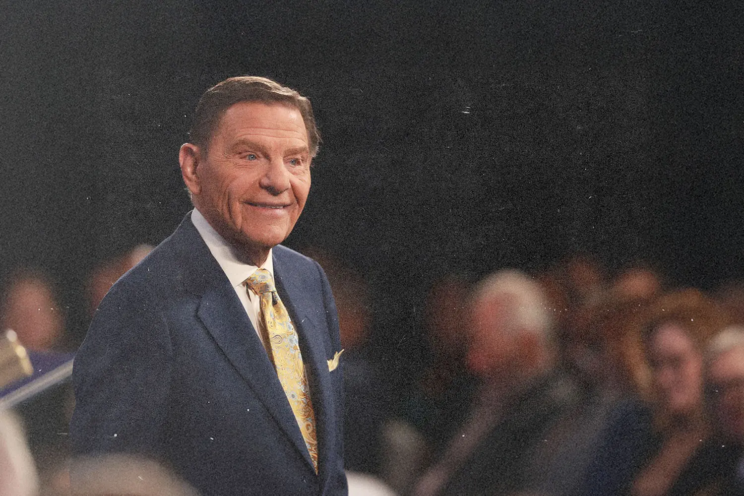 Kenneth Copeland wearing a suit and tie standing in front of an audience in a dimly lit auditorium