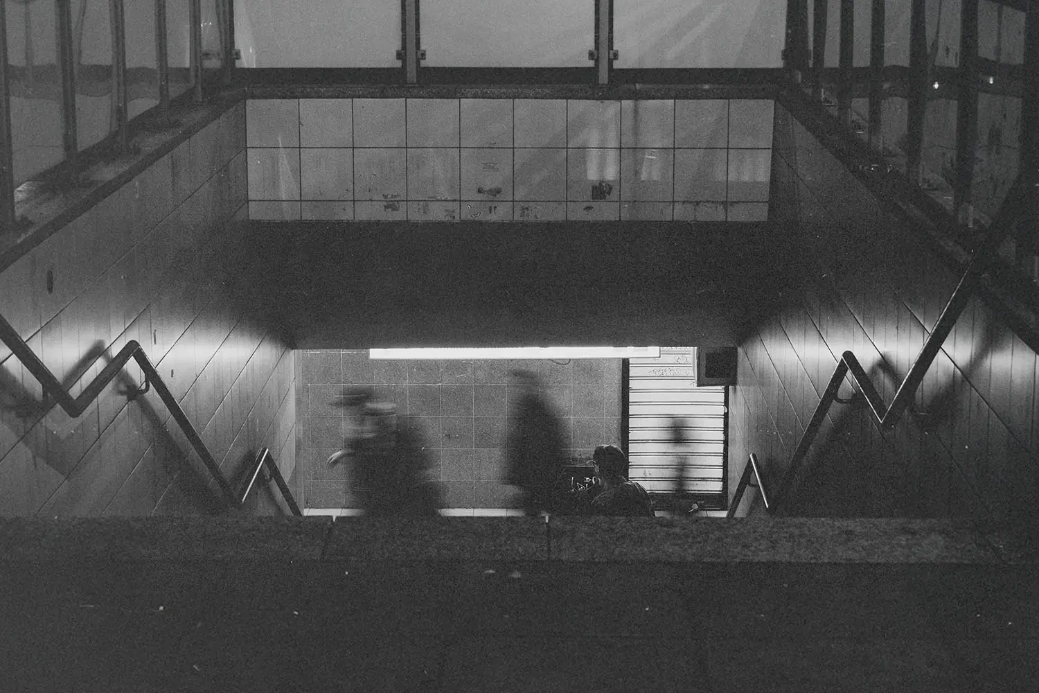 Dimly lit subway entrance with blurred figures descending toward the platform and tiled walls along the stairs