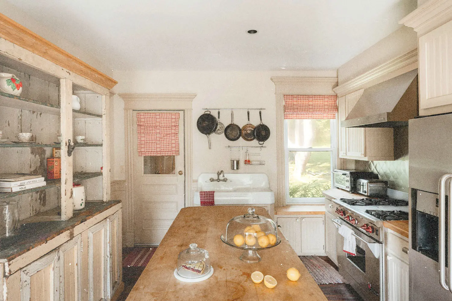 Vintage‑style kitchen with worn wooden cabinetry, hanging pans, red‑checked curtains, and lemons on a center island