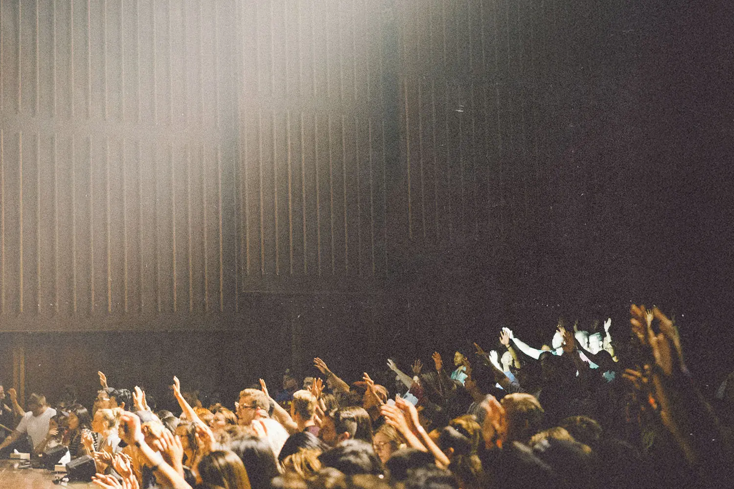 Large crowd with raised hands in a dimly lit auditorium as bright stage light shines from above