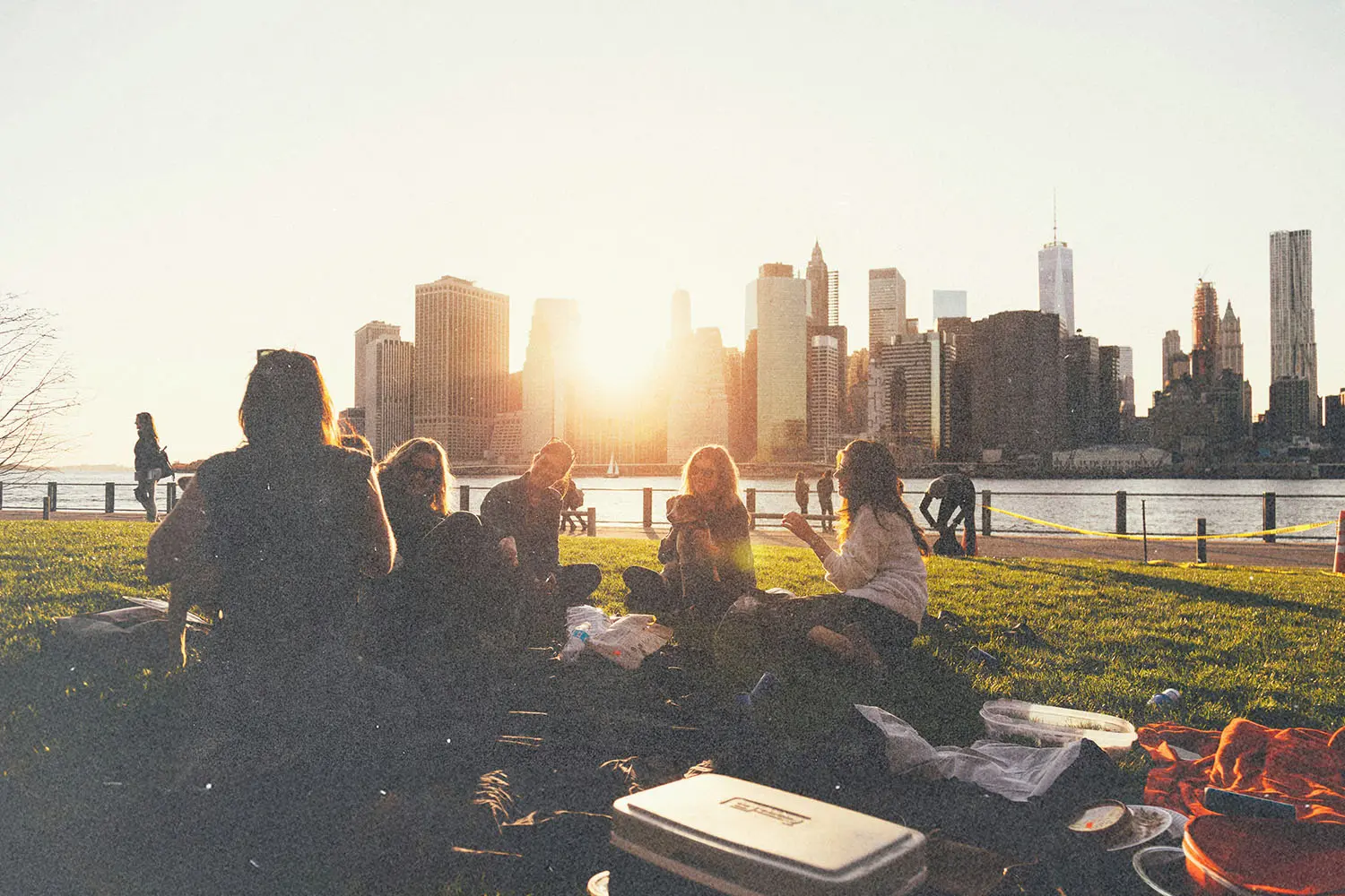 Group of people having a picnic on a grassy waterfront with a city skyline and sunset in the background