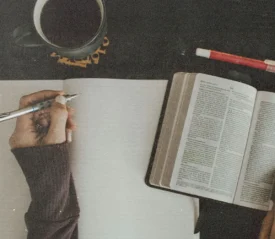 Open Bible and blank notebook on a table with a hand writing beside a cup of coffee