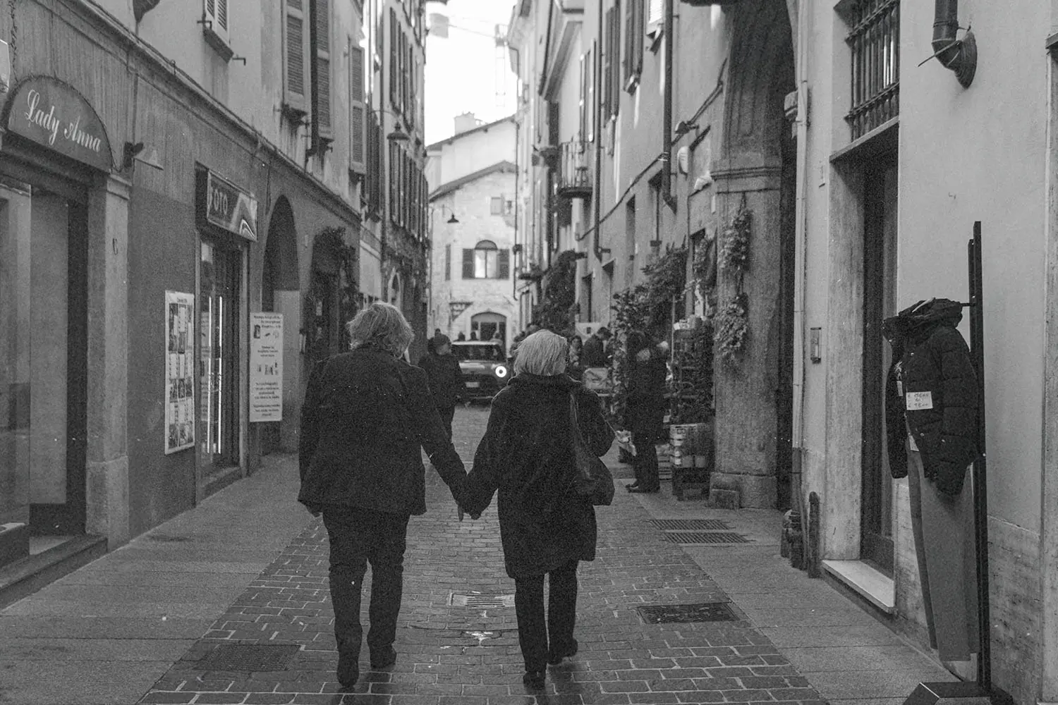 Two people walking hand‑in‑hand down a narrow cobblestone street lined with shops and tall historic buildings